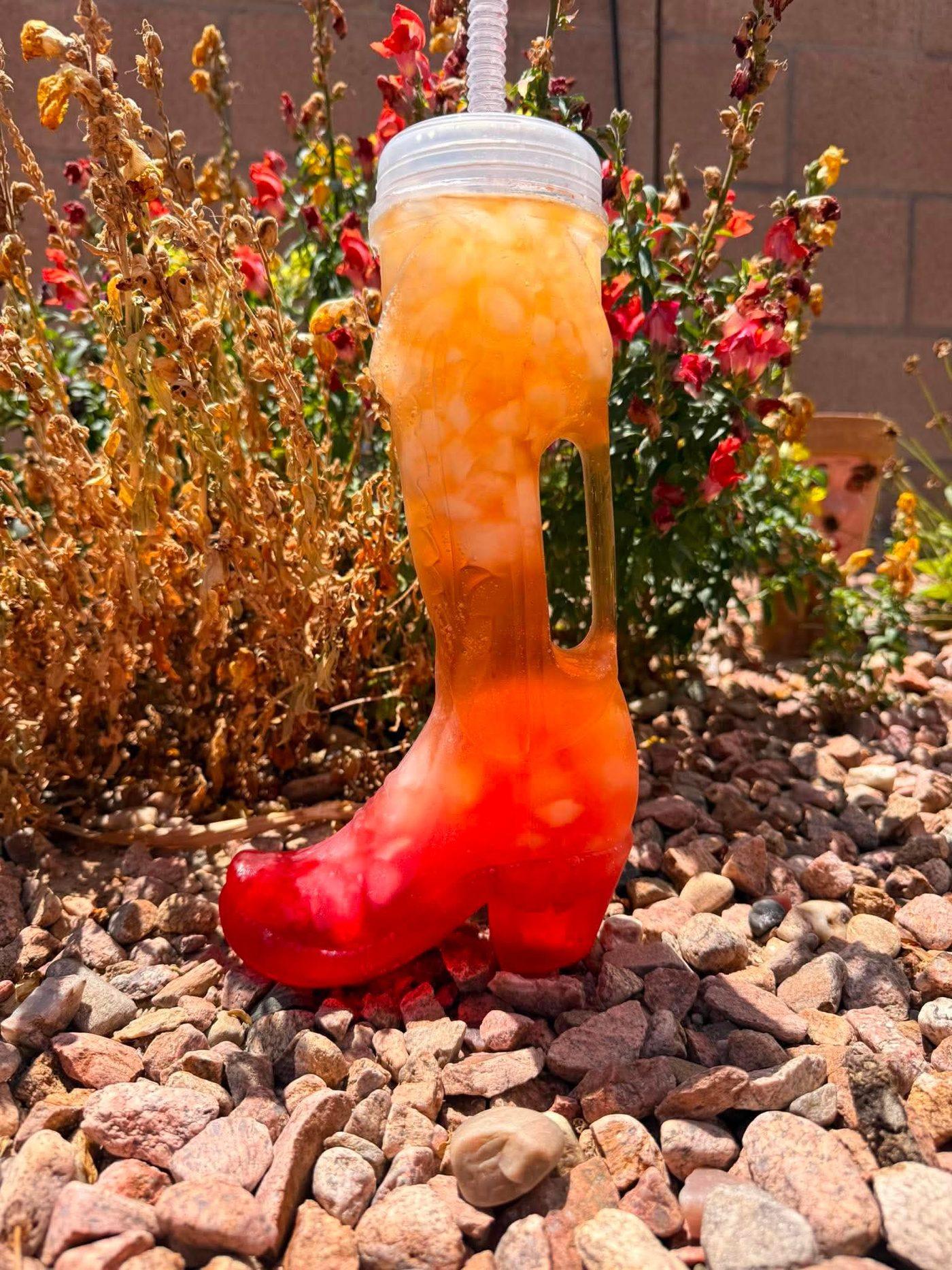 A cowboy boot cup filled with an orange and red ombre soda, sitting outdoors near flowers