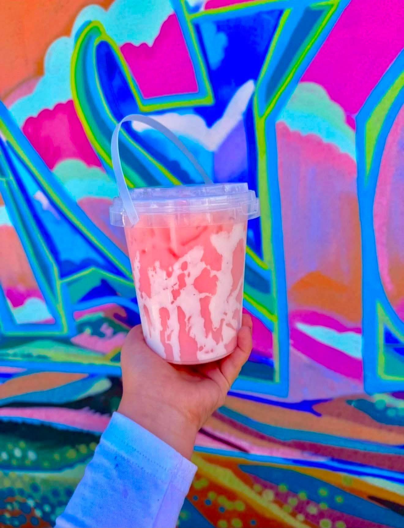 Pink swirled dirty soda in a bucket-style cup held in front of a colorful mural