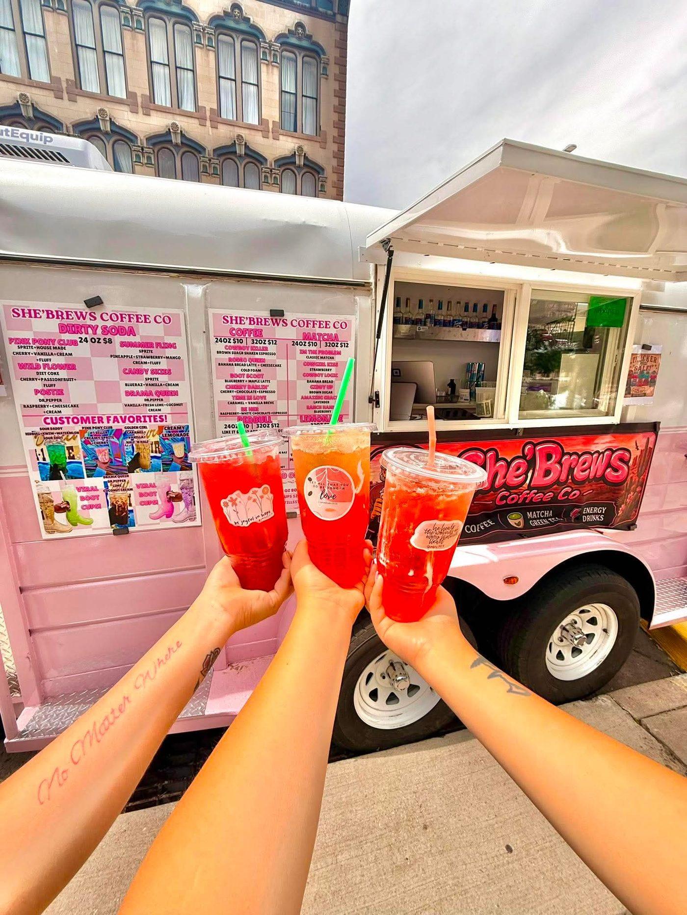 The pink She'Brews Coffee Co. trailer with menu boards, parked downtown, with three hands holding red dirty sodas