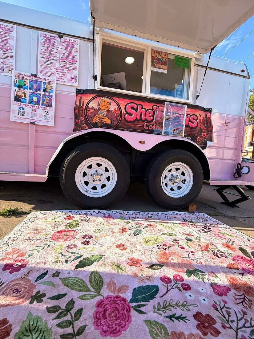 The She'Brews trailer parked with its menu boards out and a floral picnic blanket spread in front